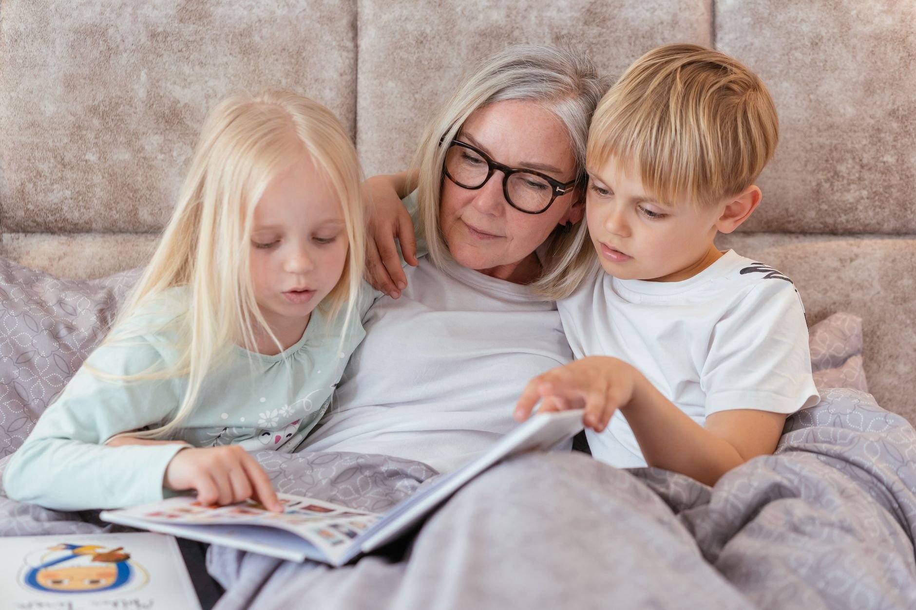 A grandmother reading a book with her grandchildren in bed, enjoying warm family time together.
